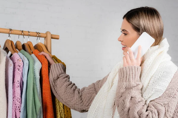 Smiling woman in scarf talking on smartphone near sweaters on hanger rack on white background — Stock Photo
