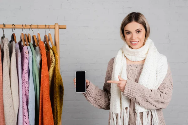 Lächelnde Frau zeigt auf Smartphone mit leerem Bildschirm neben Kleiderständer mit Pullovern auf weißem Hintergrund — Stockfoto