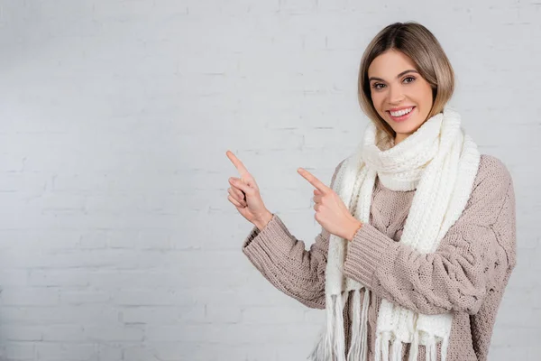 Femme souriante en vêtements chauds pointant avec les doigts sur le mur de briques blanches — Photo de stock