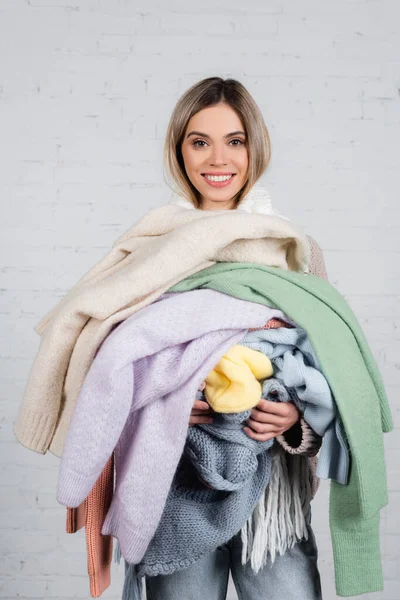 Donna sorridente che tiene maglioni lavorati a maglia e guarda la fotocamera su sfondo bianco — Foto stock