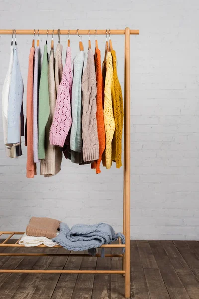Wooden hanger rack with colorful knitted sweaters near white brick wall — Stock Photo