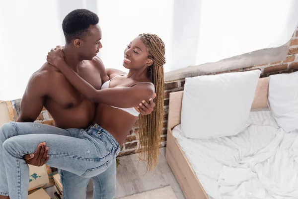 Muscular african american man holding on hands cheerful girlfriend in jeans and bra in bedroom — Stock Photo