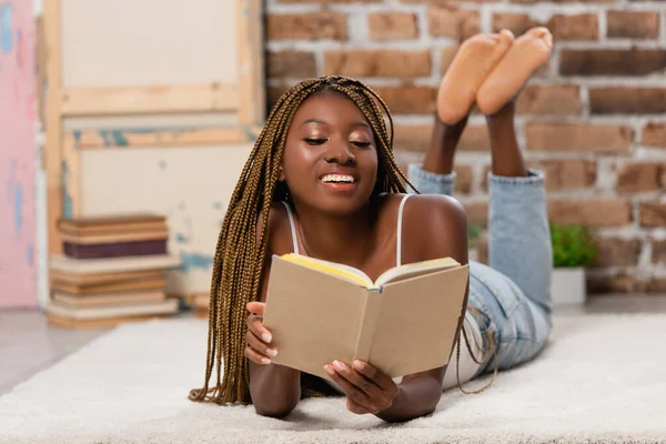 Sorrindo afro-americana leitura livro enquanto deitado no tapete em casa — Fotografia de Stock