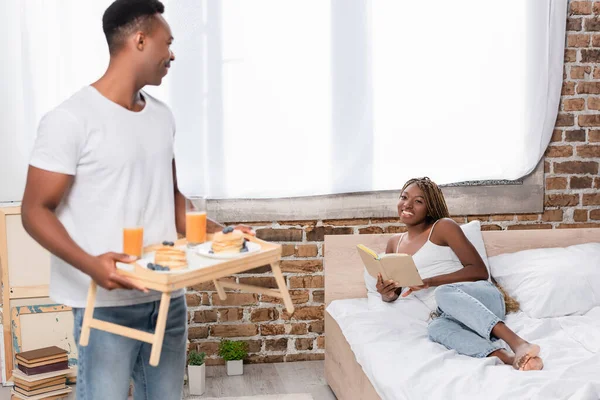 Femme afro-américaine souriante avec livre regardant petit ami avec petit déjeuner sur plateau au premier plan flou dans la chambre — Photo de stock