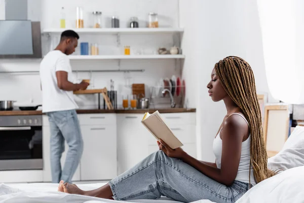 Africano americano mulher ler livro enquanto sentado na cama perto namorado na cozinha — Fotografia de Stock