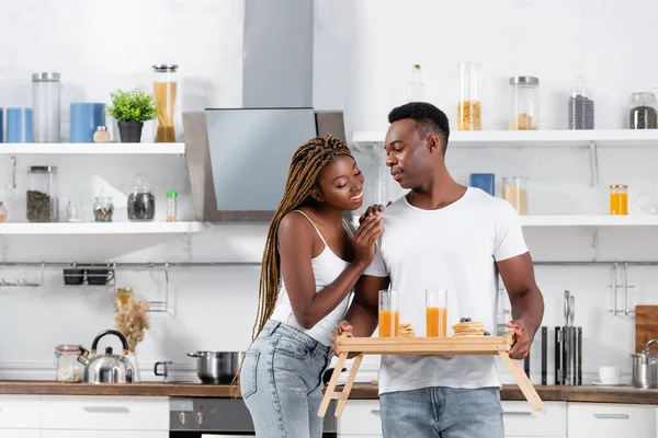 Sonriente mujer afroamericana abrazando novio con delicioso desayuno y jugo de naranja en bandeja en la cocina - foto de stock