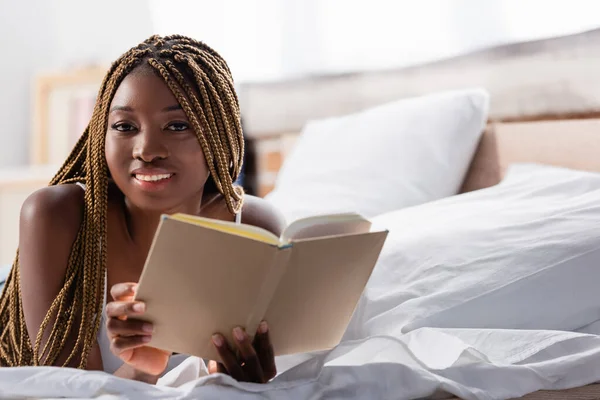 Africano americano mulher sorrindo para a câmera enquanto segurando livro em primeiro plano desfocado na cama — Fotografia de Stock