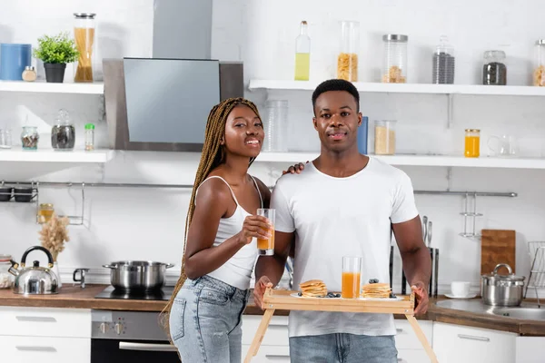 Sonriente pareja afroamericana con sabroso desayuno en bandeja y jugo de naranja mirando a la cámara en la cocina - foto de stock