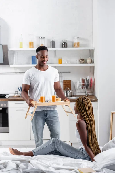 Sonriente hombre afroamericano sosteniendo jugo de naranja y panqueques en bandeja cerca de la novia en la cama en primer plano borrosa - foto de stock