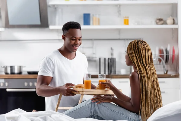 Sonriente hombre afroamericano sosteniendo bandeja con tortitas y jugo de naranja cerca de la novia en la cama durante la mañana - foto de stock