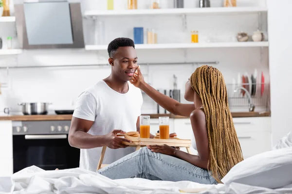 African american woman touching face of boyfriend with breakfast on tray on bed — Stock Photo