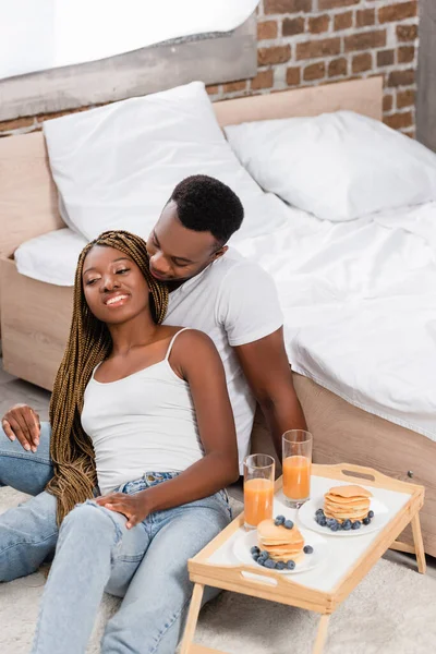 Fröhliches afrikanisch-amerikanisches Paar sitzt auf dem Boden neben dem Frühstück auf Tablett im Schlafzimmer — Stockfoto