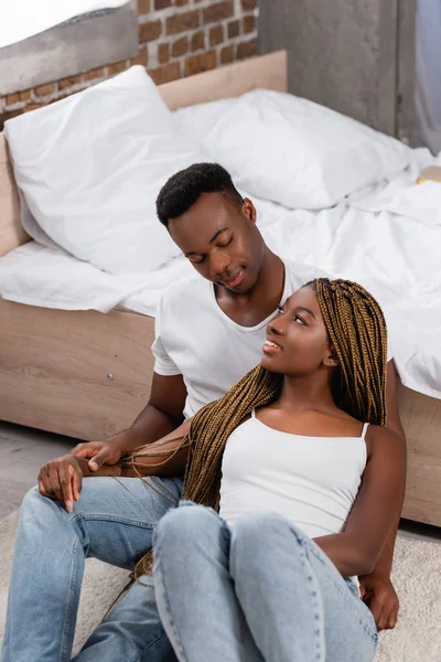 Sorrindo afro-americana olhando para o namorado perto da cama no fundo borrado — Fotografia de Stock