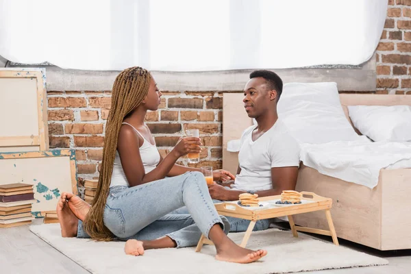 Africano casal americano em jeans segurando copos de suco de laranja perto de panquecas na bandeja no chão no quarto — Fotografia de Stock