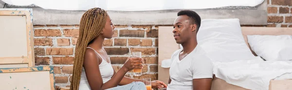 Pareja afroamericana sosteniendo vasos de jugo de naranja en el dormitorio, pancarta - foto de stock