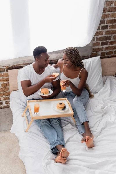 Vista de ángulo alto de la sonriente mujer afroamericana con vaso de jugo de naranja cerca de novio con panqueque en la cama - foto de stock