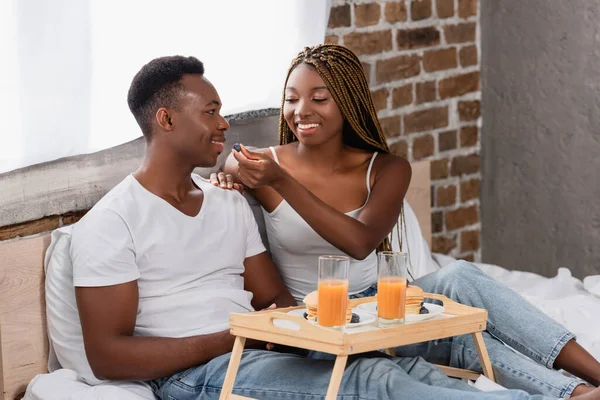 Mujer sonriente alimentando novio afroamericano con grasa cerca del desayuno en bandeja en la cama - foto de stock
