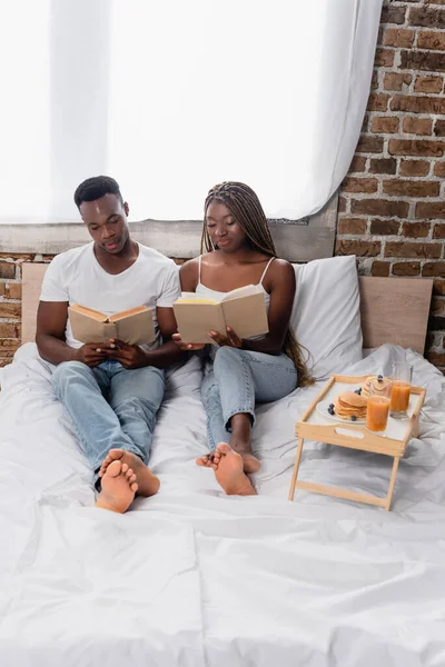 Pareja afroamericana leyendo libros cerca del desayuno en bandeja en la cama - foto de stock