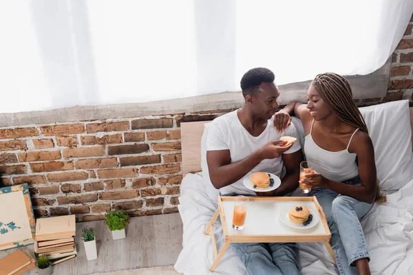 Vista de ángulo alto de la sonriente pareja afroamericana sentada en la cama cerca de delicioso desayuno en la cama - foto de stock
