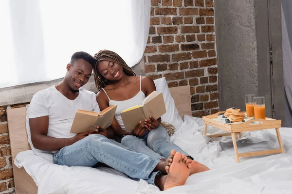 Lächelndes afrikanisches amerikanisches Paar liest Bücher in der Nähe des Frühstücks auf Tablett im Schlafzimmer — Stockfoto