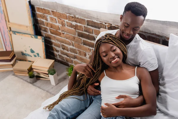 Vista de ángulo alto del hombre afroamericano sonriente abrazando a su novia con los ojos cerrados en la cama - foto de stock