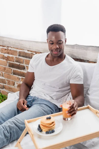 Sonriente hombre afroamericano sosteniendo un vaso de jugo de naranja cerca de panqueques en bandeja en la cama - foto de stock