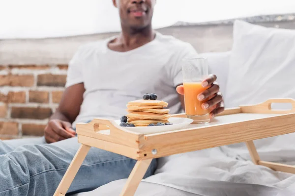 Vue recadrée de l'homme afro-américain tenant un verre de jus d'orange près de délicieuses crêpes sur plateau au lit sur fond flou — Photo de stock