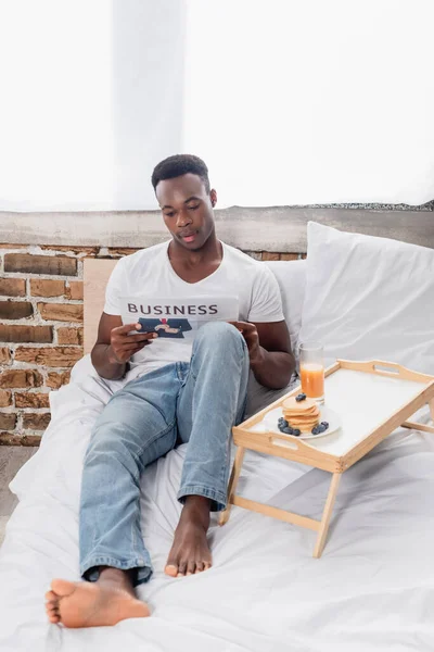 Uomo afroamericano che legge giornali d'affari vicino a frittelle e succo d'arancia sul letto — Foto stock