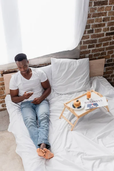 Vista de alto ângulo do homem americano africano usando smartphone perto do café da manhã e jornal de negócios na bandeja na cama — Fotografia de Stock