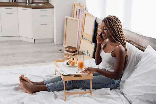 Smiling african american woman talking on smartphone near newspaper and breakfast on tray on bed — Stock Photo