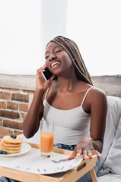 Alegre mujer afroamericana hablando en el teléfono inteligente cerca del desayuno y periódico en bandeja en la cama - foto de stock