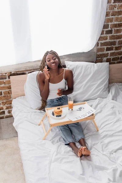 Fröhliche afrikanisch-amerikanische Frau spricht auf Smartphone neben Frühstück und Reisezeitung im Bett — Stockfoto
