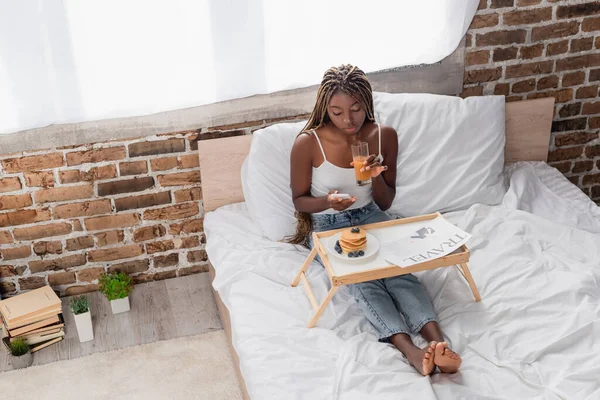 Mujer afroamericana usando smartphone y sosteniendo un vaso de jugo de naranja cerca de periódico de viaje y panqueques en la cama - foto de stock
