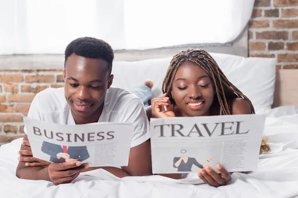 Улыбающаяся африканская американская пара читает газеты на кровати — стоковое фото