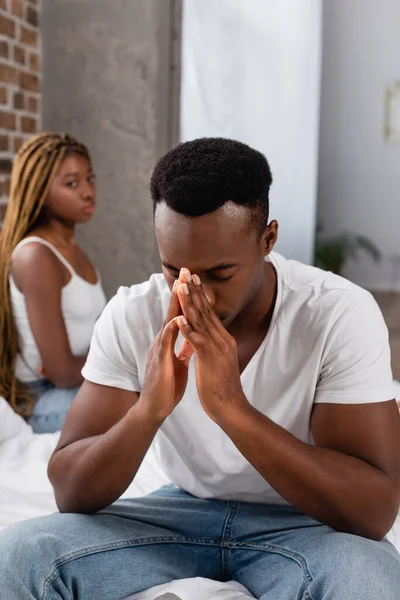 Depressed african american man sitting near girlfriend on blurred background in bedroom — Stock Photo
