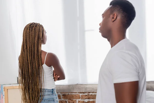 Ofendido afro-americano mulher olhando para janela perto namorado em primeiro plano desfocado em casa — Fotografia de Stock
