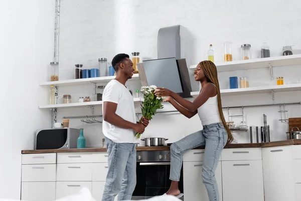 Homme afro-américain joyeux donnant des fleurs à la petite amie dans la cuisine — Photo de stock