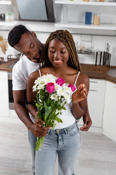 Uomo afroamericano che presenta bouquet alla fidanzata sorridente in cucina — Foto stock