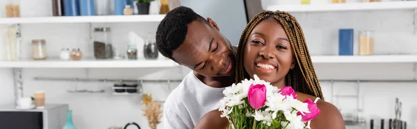 Sonriente mujer afroamericana sosteniendo ramo cerca de novio en la cocina, pancarta - foto de stock