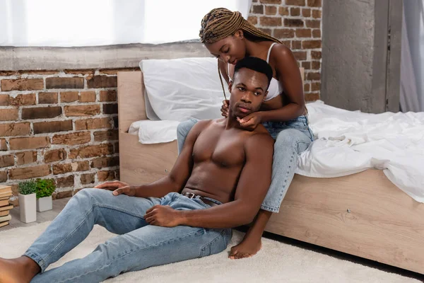 Африканська американка в джинсах і бюстгальтерах торкається безсоромного чоловіка на підлозі в спальні. — стокове фото