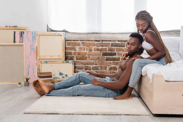 Seductive african american woman in bra touching muscular boyfriend sitting on carpet — Stock Photo