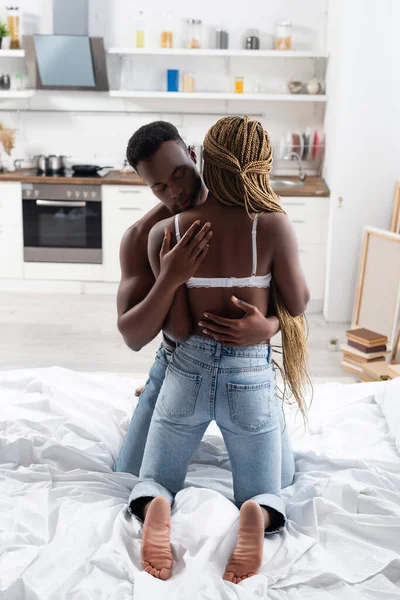 Мускулистый африканский американец обнимает девушку в лифчике и джинсах на кровати — стоковое фото