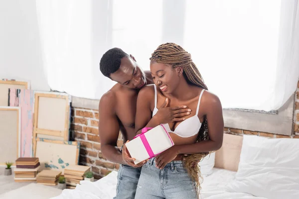 Sexy africano americano uomo dare regalo per allegro ragazza in reggiseno in camera da letto — Foto stock