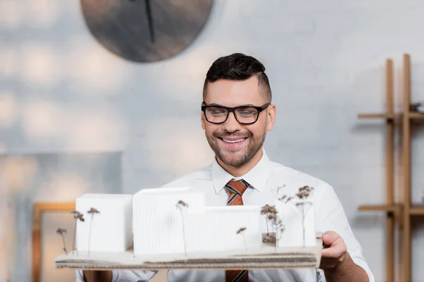 Cheerful architect in eyeglasses holding buildings maquette in architectural agency — Stock Photo