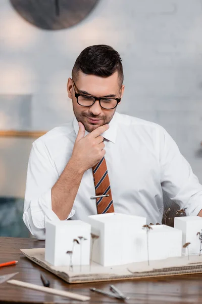 Thoughtful, positive architect looking at house models in office — Stock Photo