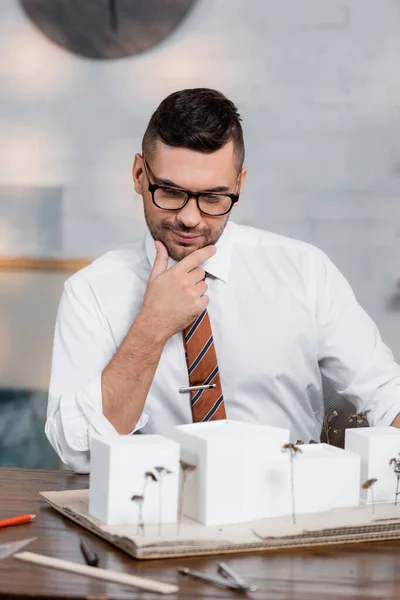 Nachdenklicher, positiver Architekt, der Hausmodelle im Büro betrachtet — Stockfoto