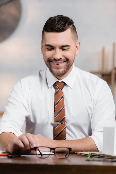 Happy architect sitting at workplace near eyeglasses on desk, blurred foreground — Stock Photo