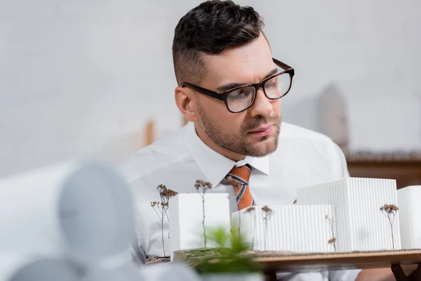 Serious architect in eyeglasses looking at buildings models in office, blurred foreground — Stock Photo