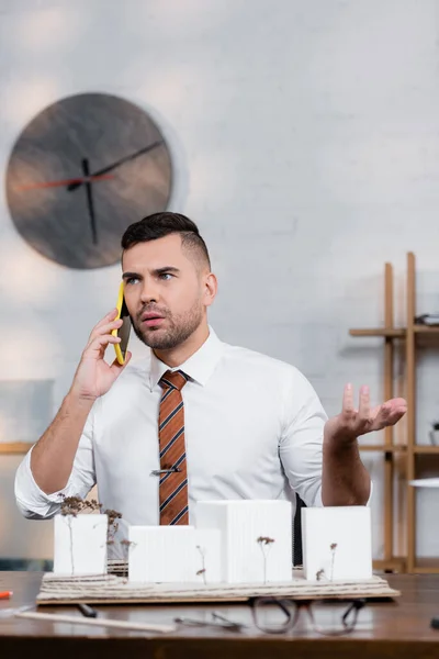 Serious architect gesturing while talking on smartphone near architectural project, blurred foreground — Stock Photo