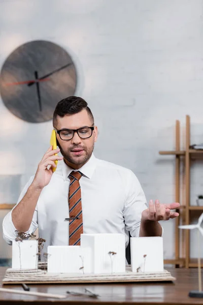 Architecte sérieux parler sur téléphone portable et geste près des modèles de maisons — Photo de stock